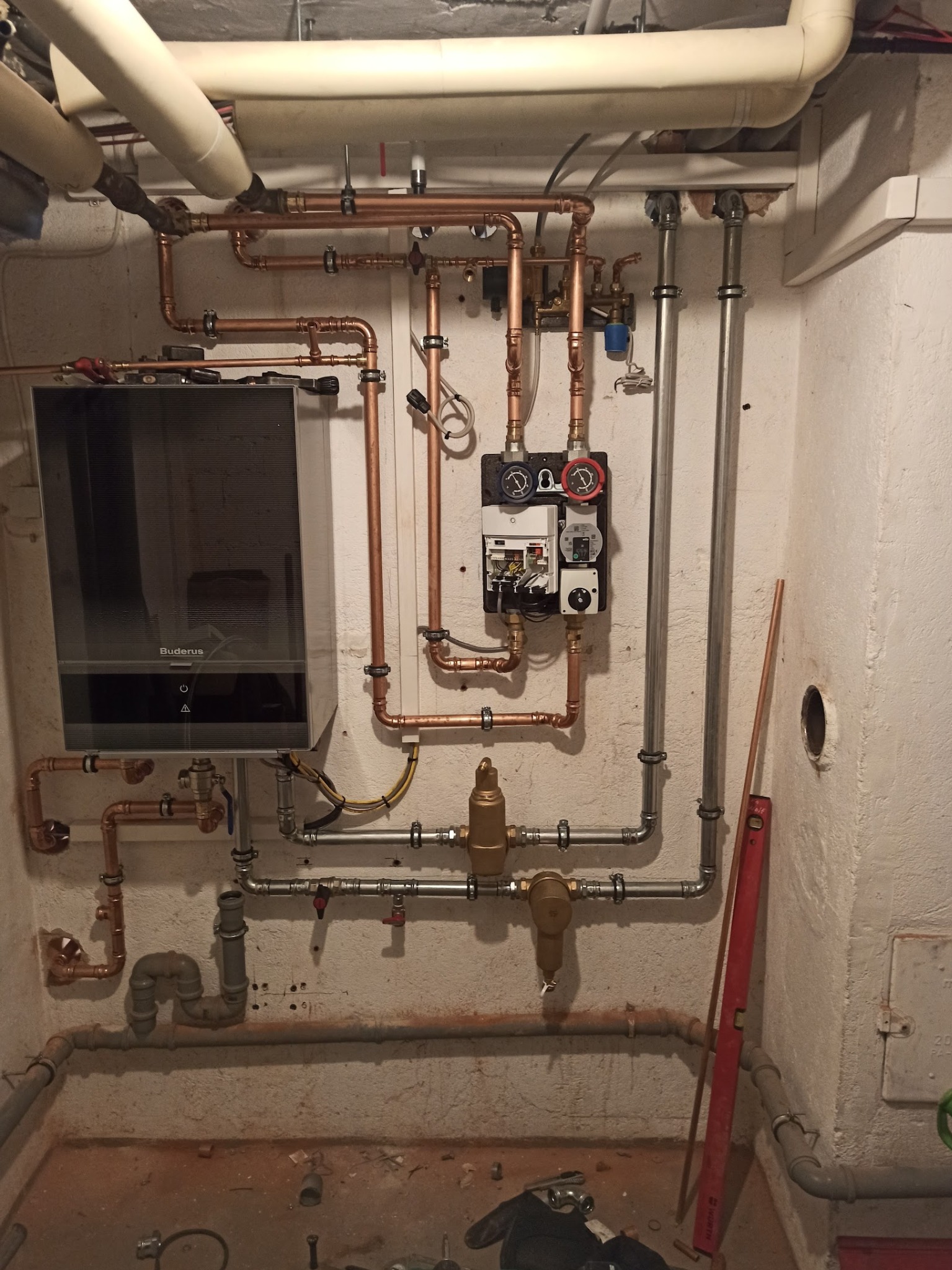 Złożona instalacja grzewcza z piecem Buderus, rurami miedzianymi i stalowymi, manometrami i zaworami, zamontowana w piwnicy o surowych ścianach.