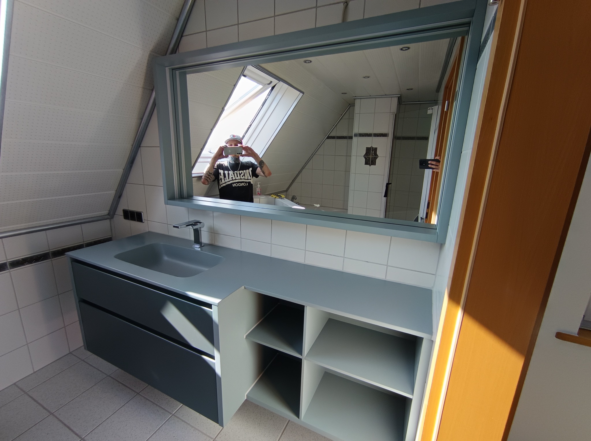 Nowoczesna szara szafka łazienkowa z umywalką i otwartymi półkami, widoczna w lustrze osoba robiąca zdjęcie w łazience na poddaszu.