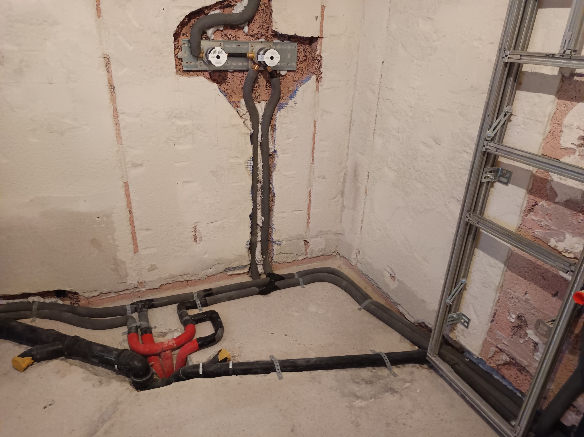 Szare i czerwone rury instalacji hydraulicznej w surowym wnętrzu, widoczne mocowania do ściany i podłogi, fragment metalowej konstrukcji w rogu pomieszczenia.