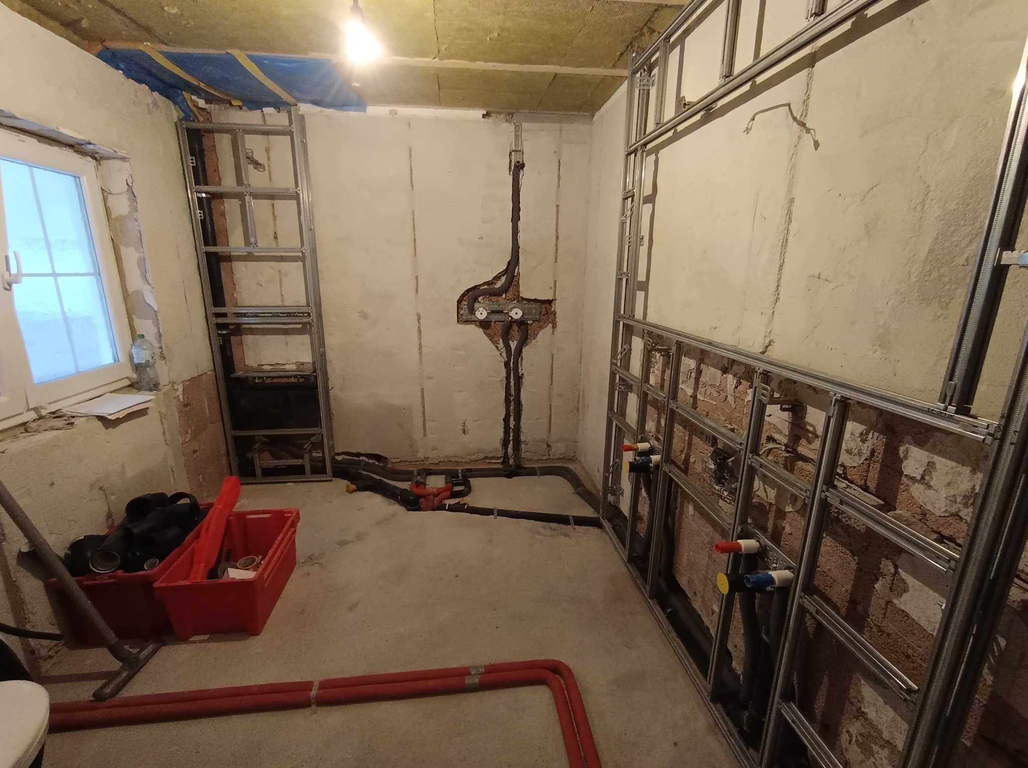 Wnętrze w trakcie instalacji hydraulicznej: widoczne rury, metalowe stelaże przy ścianach, narzędzia w czerwonym pojemniku. Surowy stan wykończenia, okno z lewej strony.