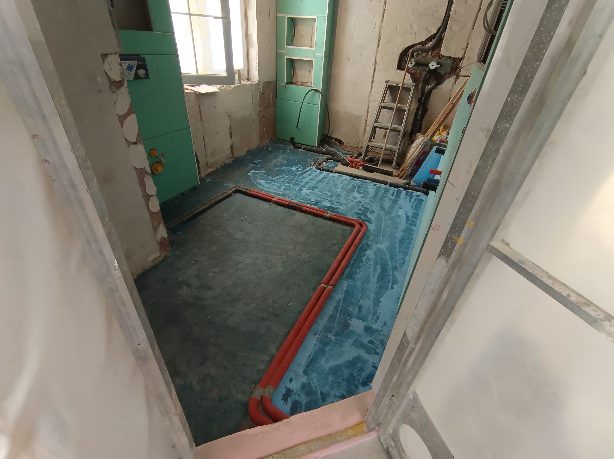 Instalacja ogrzewania podłogowego w trakcie remontu: widoczne rury grzewcze na izolacji, ściany w surowym stanie, drabina i narzędzia w tle. Etap prac przed wylaniem posadzki.