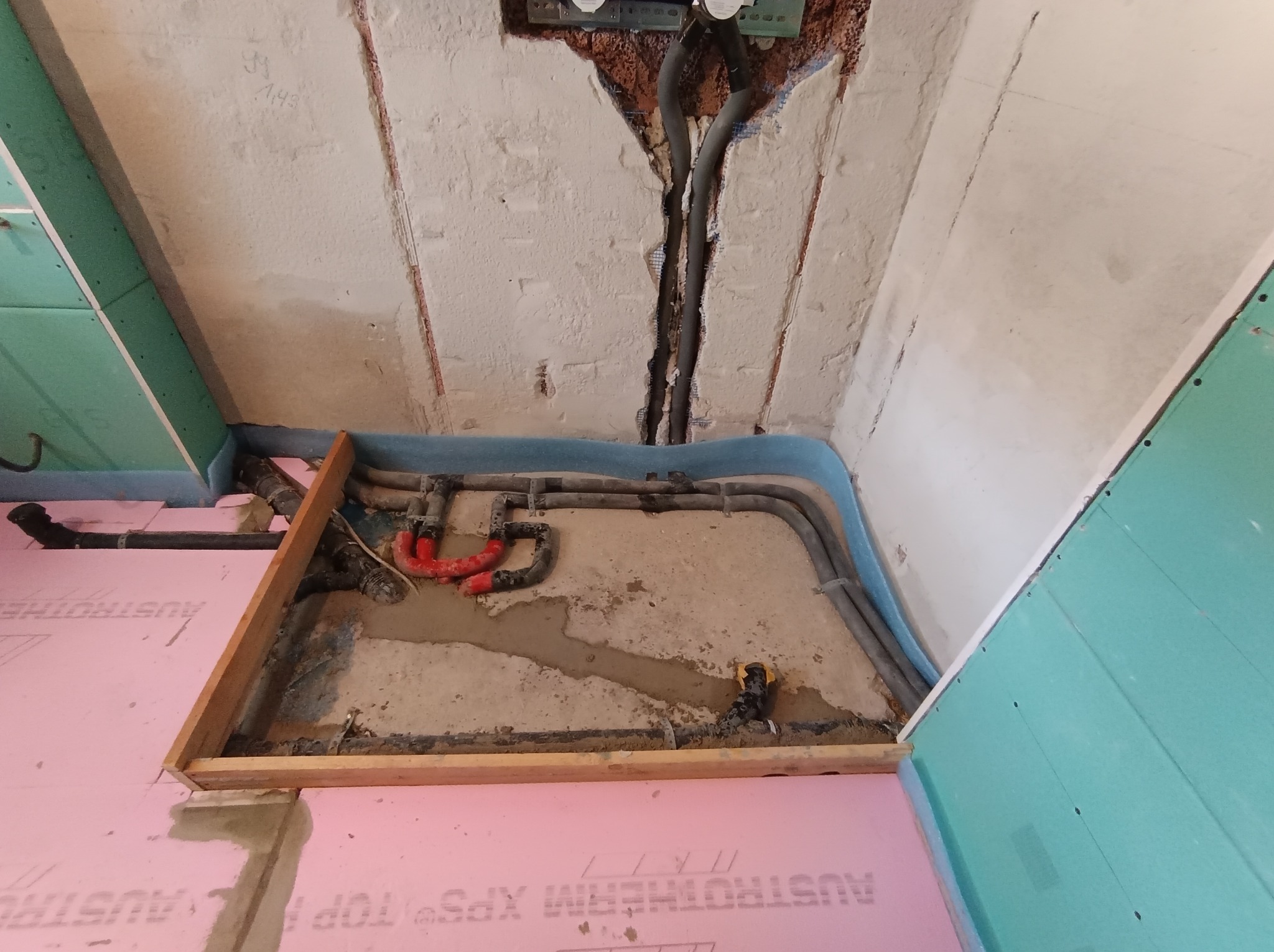 Fragment instalacji ogrzewania podłogowego w trakcie montażu: rury grzewcze ułożone na izolacji, widoczne przyłącza i elementy mocujące. Surowy stan wykończenia ścian.