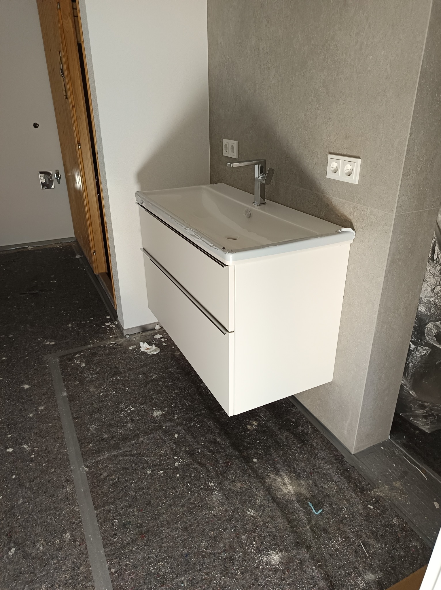 Biała, podwieszana szafka łazienkowa z umywalką i chromowaną baterią na tle szarej ściany z gniazdkami. Widoczne ślady prac wykończeniowych na podłodze.
