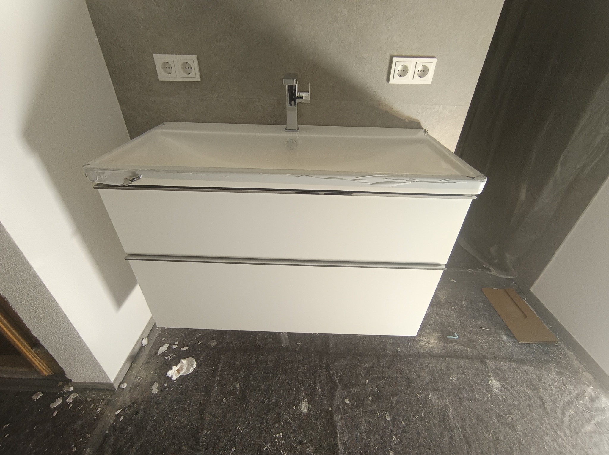 Nowoczesna biała szafka pod umywalkę z chromowaną baterią, zabezpieczona folią ochronną. Widoczne gniazdka elektryczne na szarej ścianie i fragmenty gruzu na podłodze.