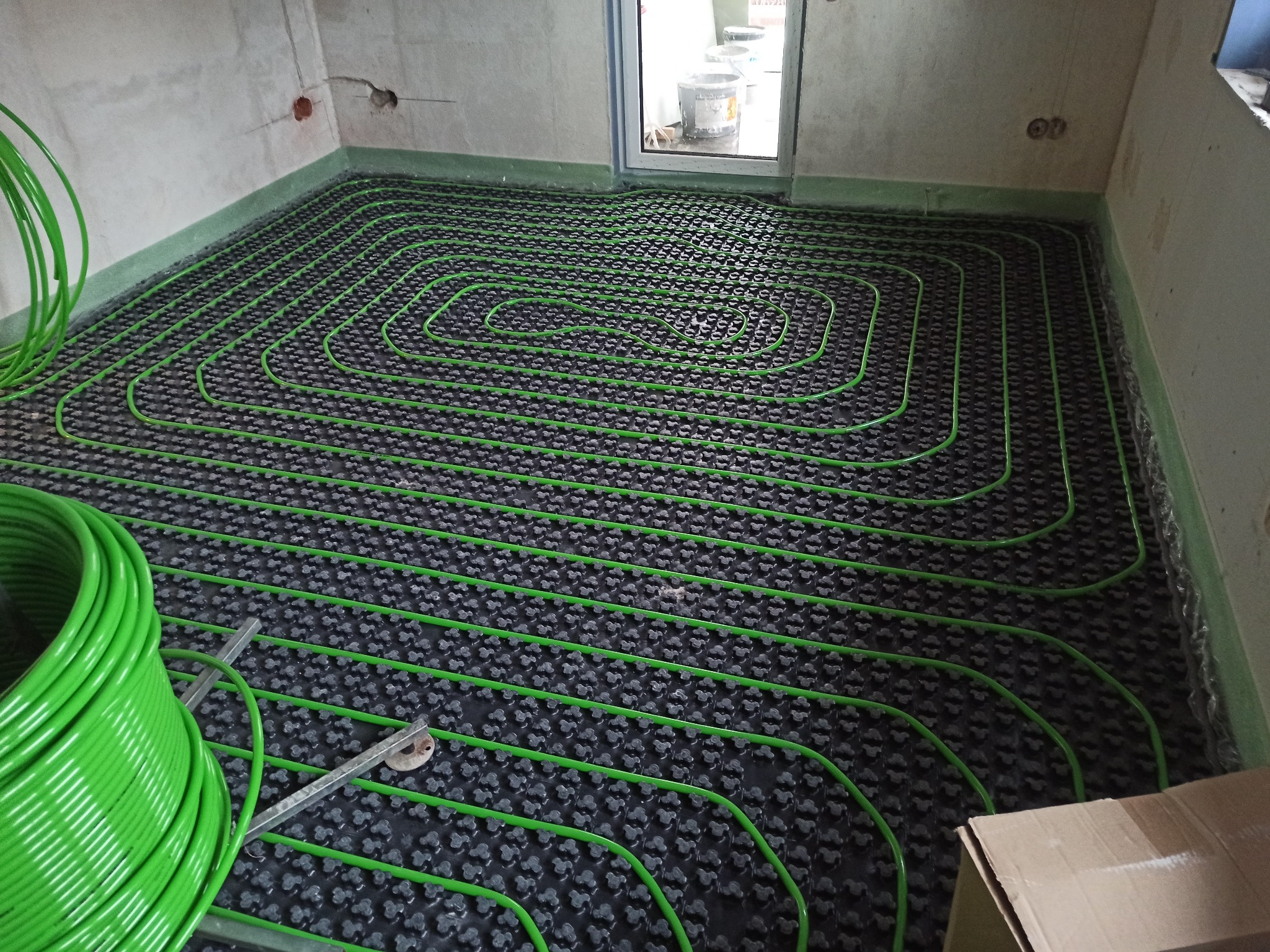 Zielone rury ogrzewania podłogowego ułożone na czarnej macie w surowym wnętrzu. Widoczna rolka rur i narzędzia. Etap instalacji przed wylewką.