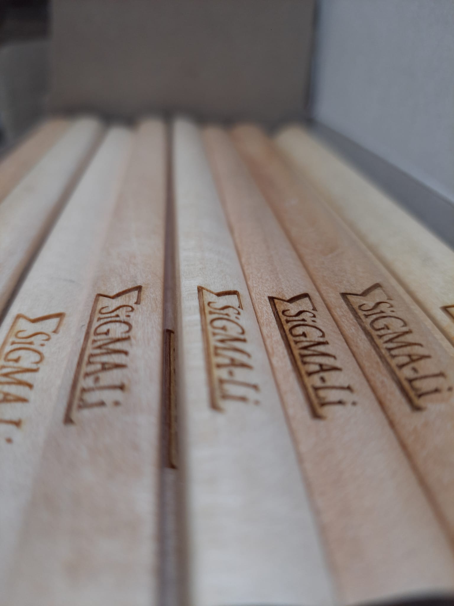 Bliski plan drewnianych listewek z wygrawerowanym logo 'SIGMA-Li', ułożonych równolegle w pudełku, z efektem głębi ostrości.