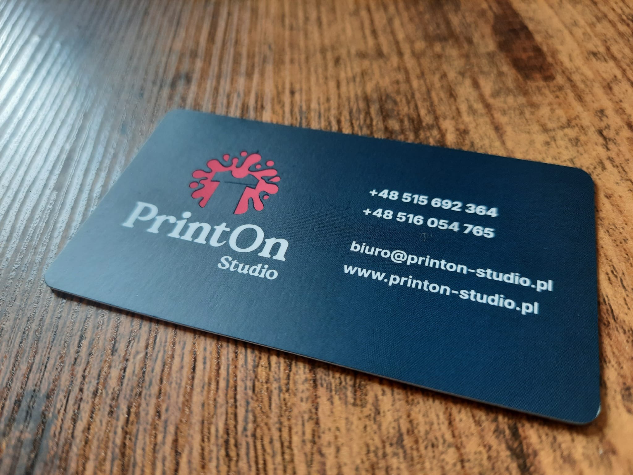 Elegancka, matowa wizytówka firmy PrintOn Studio w kolorze granatowym z czerwonym logo i białymi danymi kontaktowymi, leżąca na drewnianym blacie. Zaokrąglone rogi.