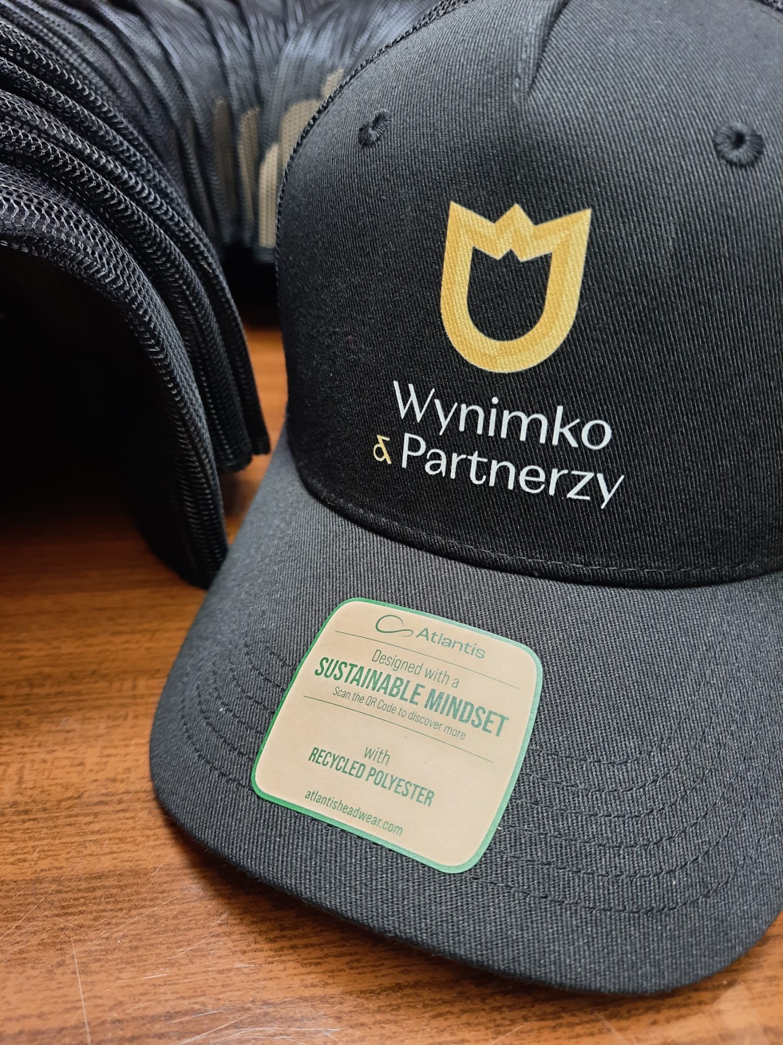 Czarna czapka z daszkiem z haftowanym logo 'Wynimko & Partnerzy' i złotym symbolem. Widoczna metka 'Sustainable Mindset' i stos czapek w tle.