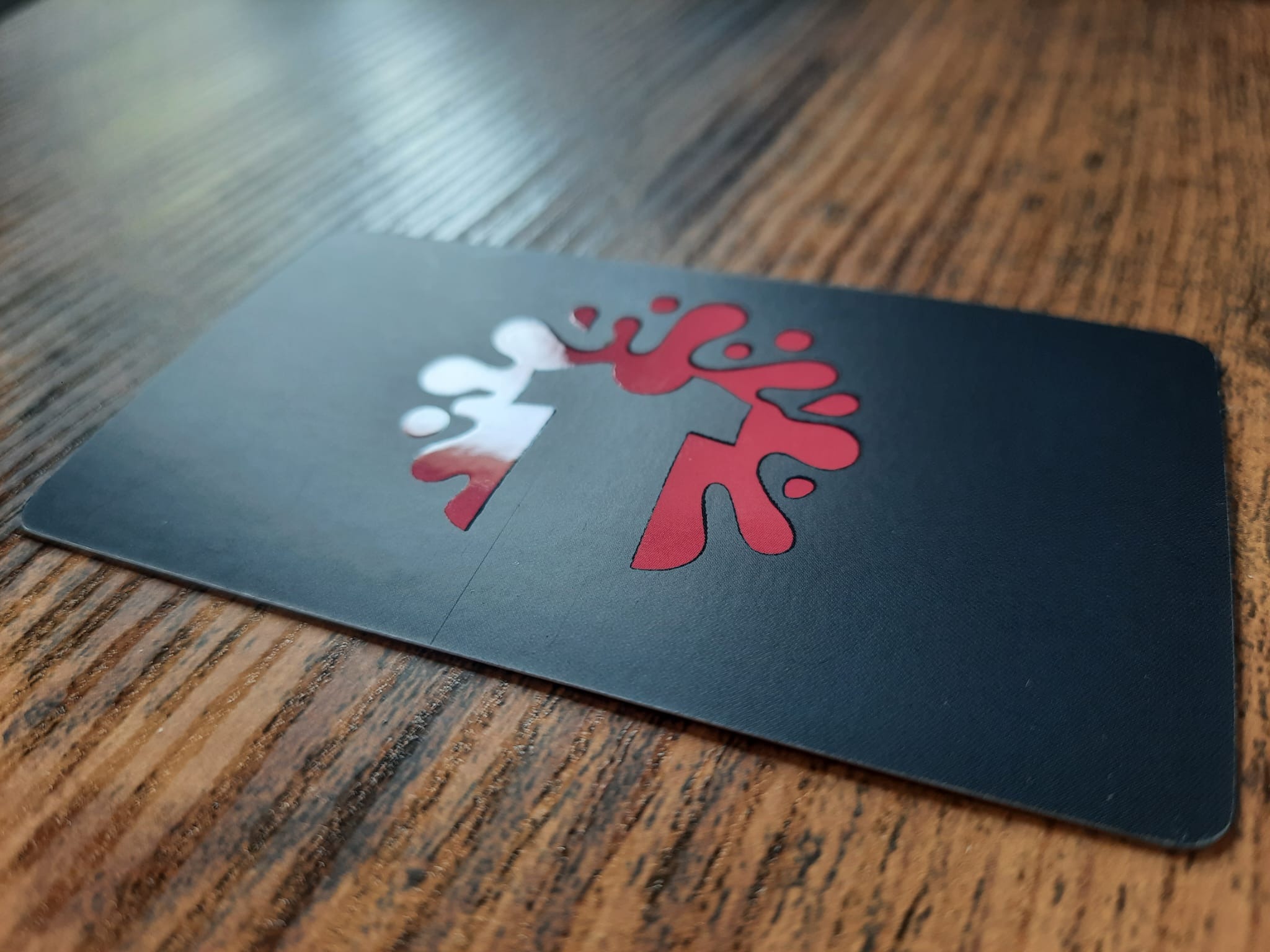 Czarna wizytówka z wyciętym, czerwono-białym logo na drewnianym tle. Minimalistyczny design, matowe wykończenie, unikalny kształt logo.