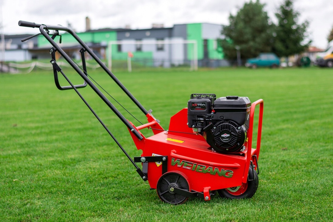 Czerwony aerator spalinowy Weibang na zielonym trawniku, w tle budynek z elementami zieleni i boisko. Sprzęt do napowietrzania gleby na trawnikach.