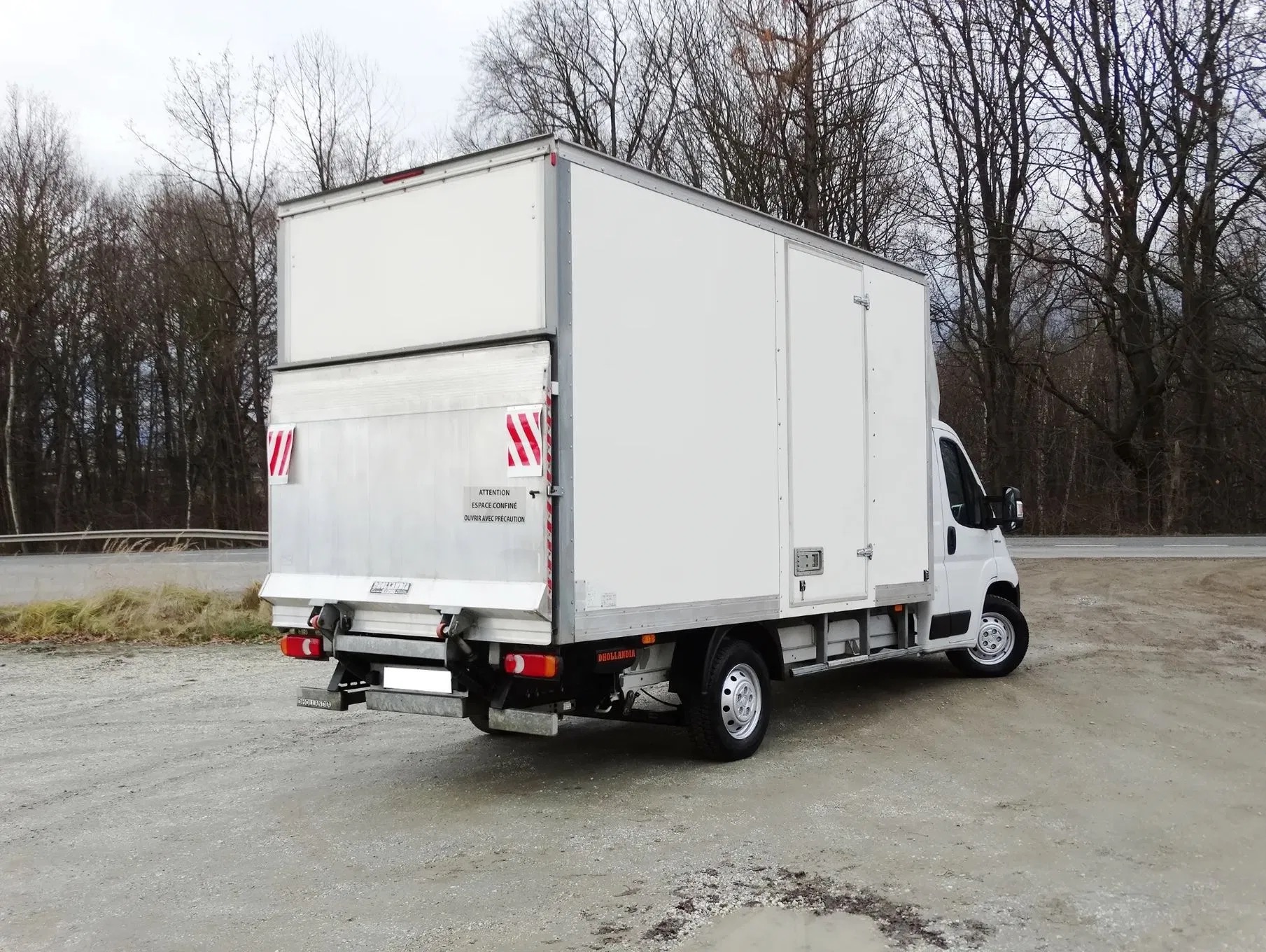 Biały bus dostawczy z otwartą przestrzenią ładunkową, zaparkowany na szutrowej nawierzchni, z widocznymi drzewami w tle. Widok z tyłu i boku.