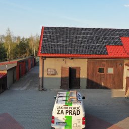 SITARSKY ENERGY SPÓŁKA Z OGRANICZONĄ ODPOWIEDZIALNOŚCIĄ - Instalacja paneli słonecznych na dachu budynku gospodarczego z czerwonym dachem. Na pierwszym planie biały samochód z napisem 'Jak nie płacić za prąd'.