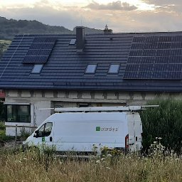 SITARSKY ENERGY SPÓŁKA Z OGRANICZONĄ ODPOWIEDZIALNOŚCIĄ - Dom z panelami słonecznymi na dachu, częściowo zasłonięty przez biały samochód dostawczy z logo firmy. W tle górzysty krajobraz i zachmurzone niebo.