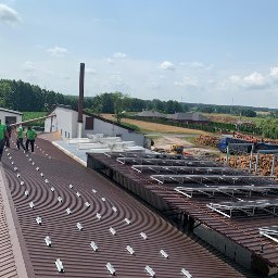 SITARSKY ENERGY SPÓŁKA Z OGRANICZONĄ ODPOWIEDZIALNOŚCIĄ - Montaż paneli fotowoltaicznych na dachu budynku przemysłowego. Widoczni pracownicy w zielonych koszulkach. W tle drzewa i zabudowania.