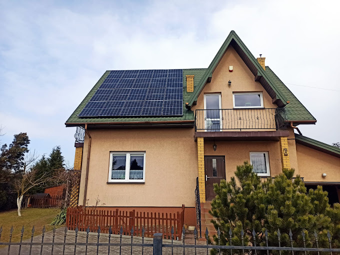 Dom jednorodzinny z panelami fotowoltaicznymi na dachu, widok z zewnątrz, ogrodzenie metalowe na pierwszym planie, pochmurne niebo w tle.