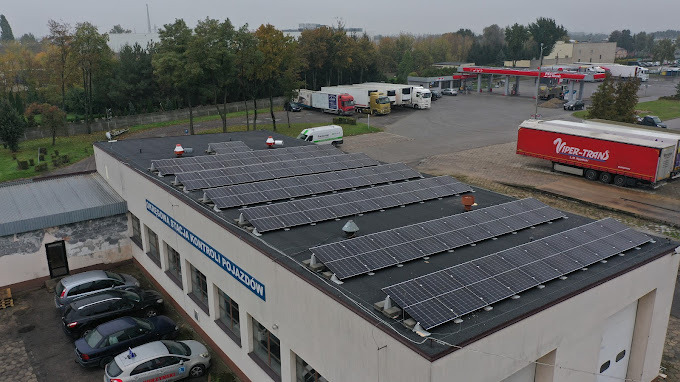 Instalacja fotowoltaiczna na dachu budynku stacji kontroli pojazdów. W tle stacja benzynowa i zaparkowane samochody ciężarowe. Pochmurny dzień.