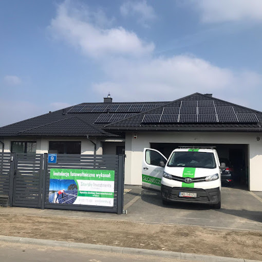 Dom jednorodzinny z panelami fotowoltaicznymi na dachu, obok zaparkowany biały samochód dostawczy z logo firmy. Na ogrodzeniu reklama instalacji fotowoltaicznej.