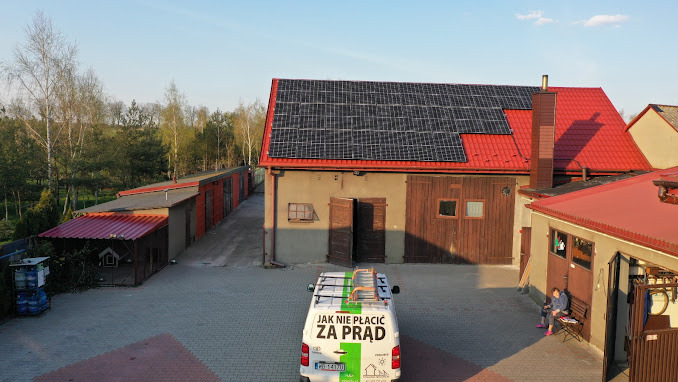 Instalacja paneli słonecznych na dachu budynku gospodarczego z czerwonym dachem. Na pierwszym planie biały samochód z napisem 'Jak nie płacić za prąd'.