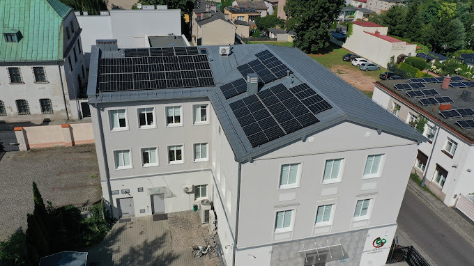 Instalacja fotowoltaiczna na dachu budynku z szarej blachy, widok z góry. Panele rozmieszczone na różnych poziomach dachu, w tle zabudowa miejska.