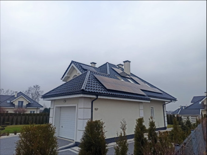 Dom z panelami słonecznymi na dachu w pochmurny dzień. Widoczne panele fotowoltaiczne na ciemnym dachu, jasna elewacja budynku i zadbany ogród. Ujęcie z dołu.