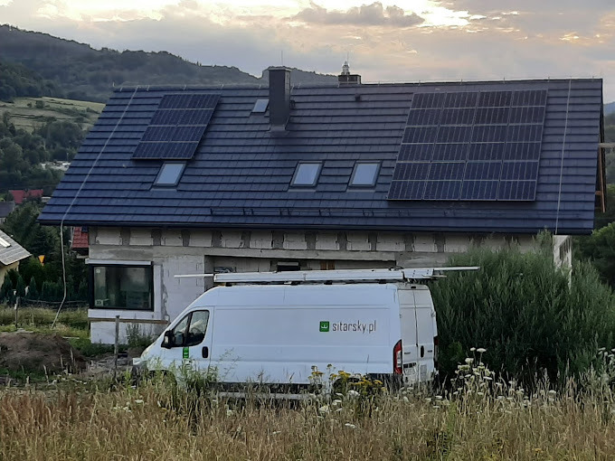 Dom z panelami słonecznymi na dachu, częściowo zasłonięty przez biały samochód dostawczy z logo firmy. W tle górzysty krajobraz i zachmurzone niebo.