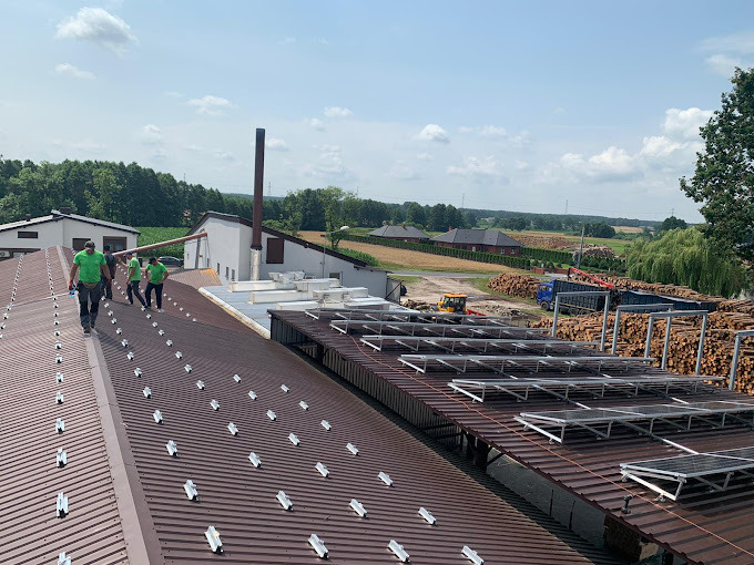 Montaż paneli fotowoltaicznych na dachu budynku przemysłowego. Widoczni pracownicy w zielonych koszulkach. W tle drzewa i zabudowania.