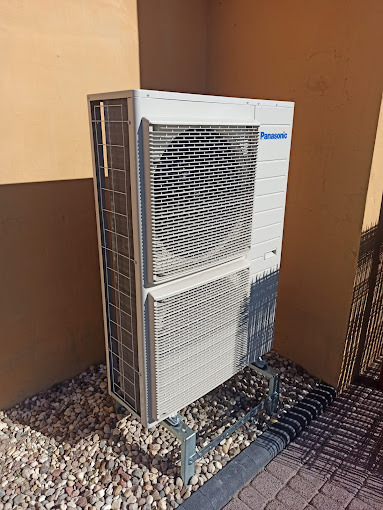 Biała pompa ciepła Panasonic, zamontowana na zewnątrz budynku na żwirowym podłożu, widoczna w narożniku ściany w słoneczny dzień.