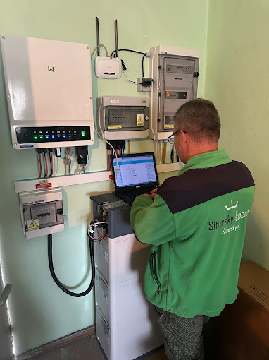 Instalator w zielonej bluzie konfiguruje system z inwerterem i magazynem energii, używając laptopa. Widoczne okablowanie i elementy instalacji elektrycznej na ścianie.
