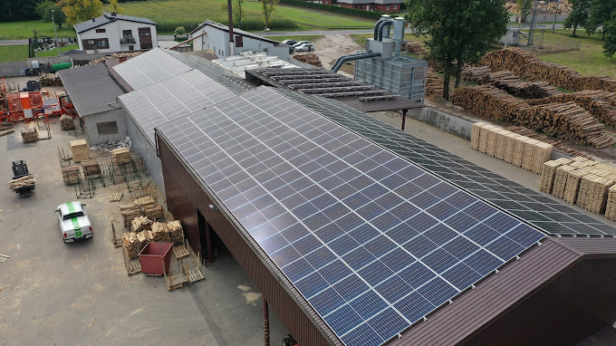Widok z góry na budynek z panelami słonecznymi na dachu. W tle składowane drewno i budynki gospodarcze. Jasne, naturalne oświetlenie.