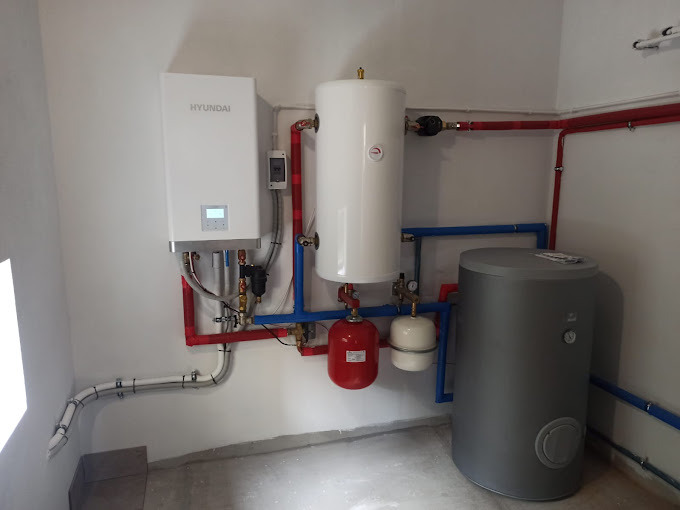 Biała pompa ciepła Hyundai z zbiornikami wyrównawczymi (czerwony i biały) i szarym zasobnikiem ciepłej wody, połączone rurami w kolorach czerwonym i niebieskim.