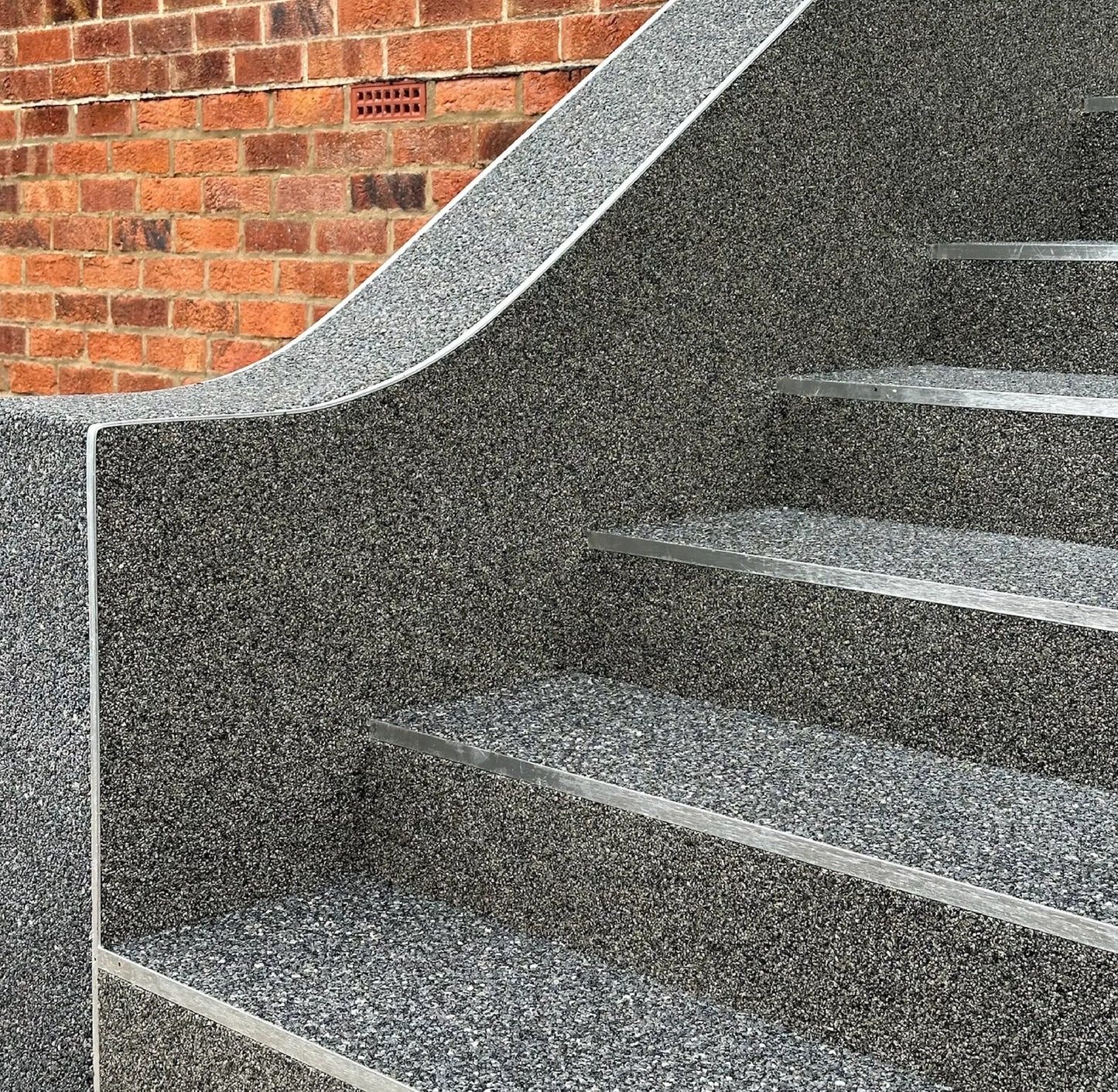 Schody zewnętrzne z szarej, drobnoziarnistej nawierzchni, wykończone metalowymi listwami, na tle ceglanej ściany. Detal wykończenia.