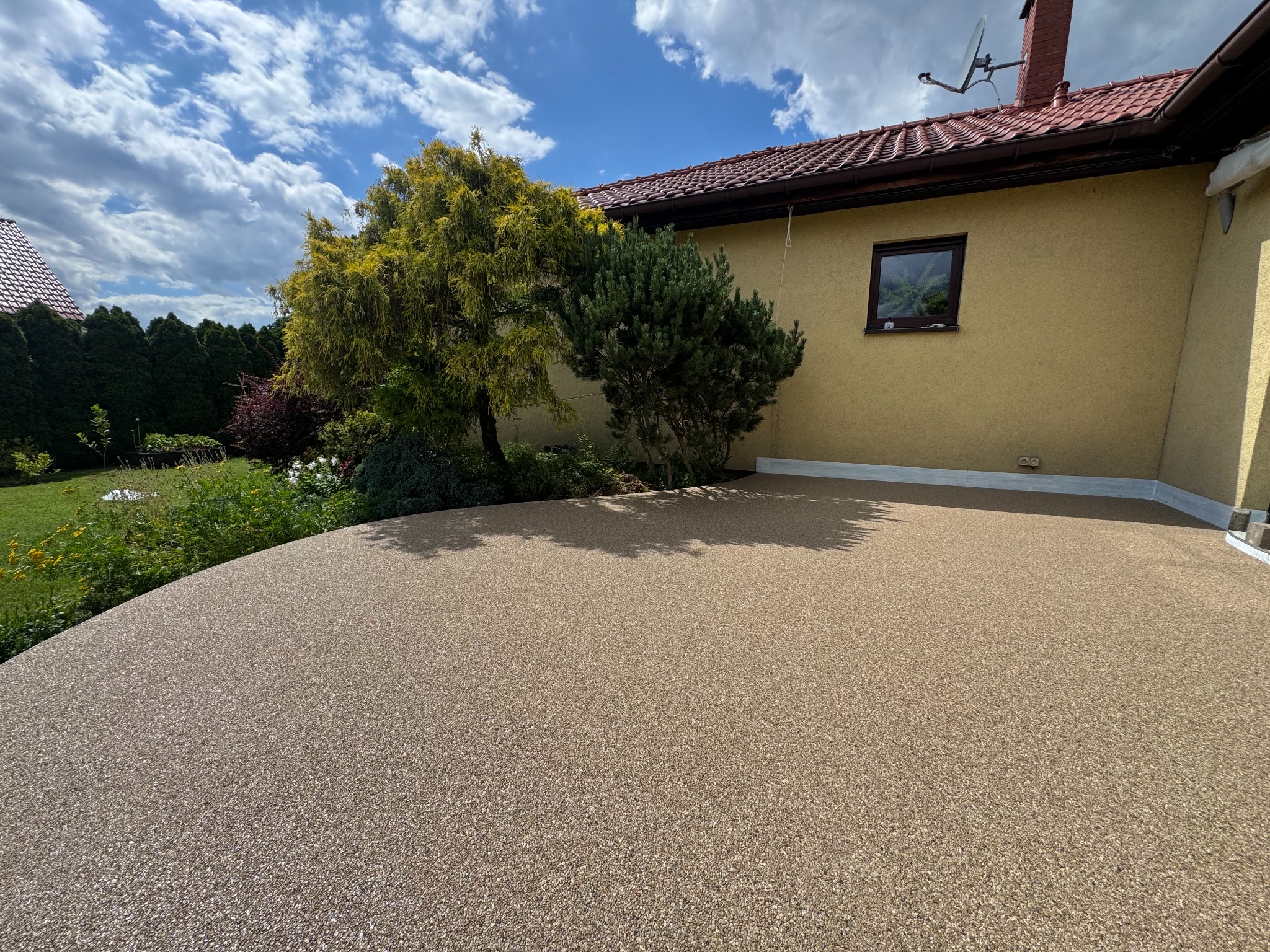 Beżowa posadzka żywiczna na zewnątrz budynku jednorodzinnego, widok z poziomu posadzki, w tle zieleń i błękitne niebo z chmurami.