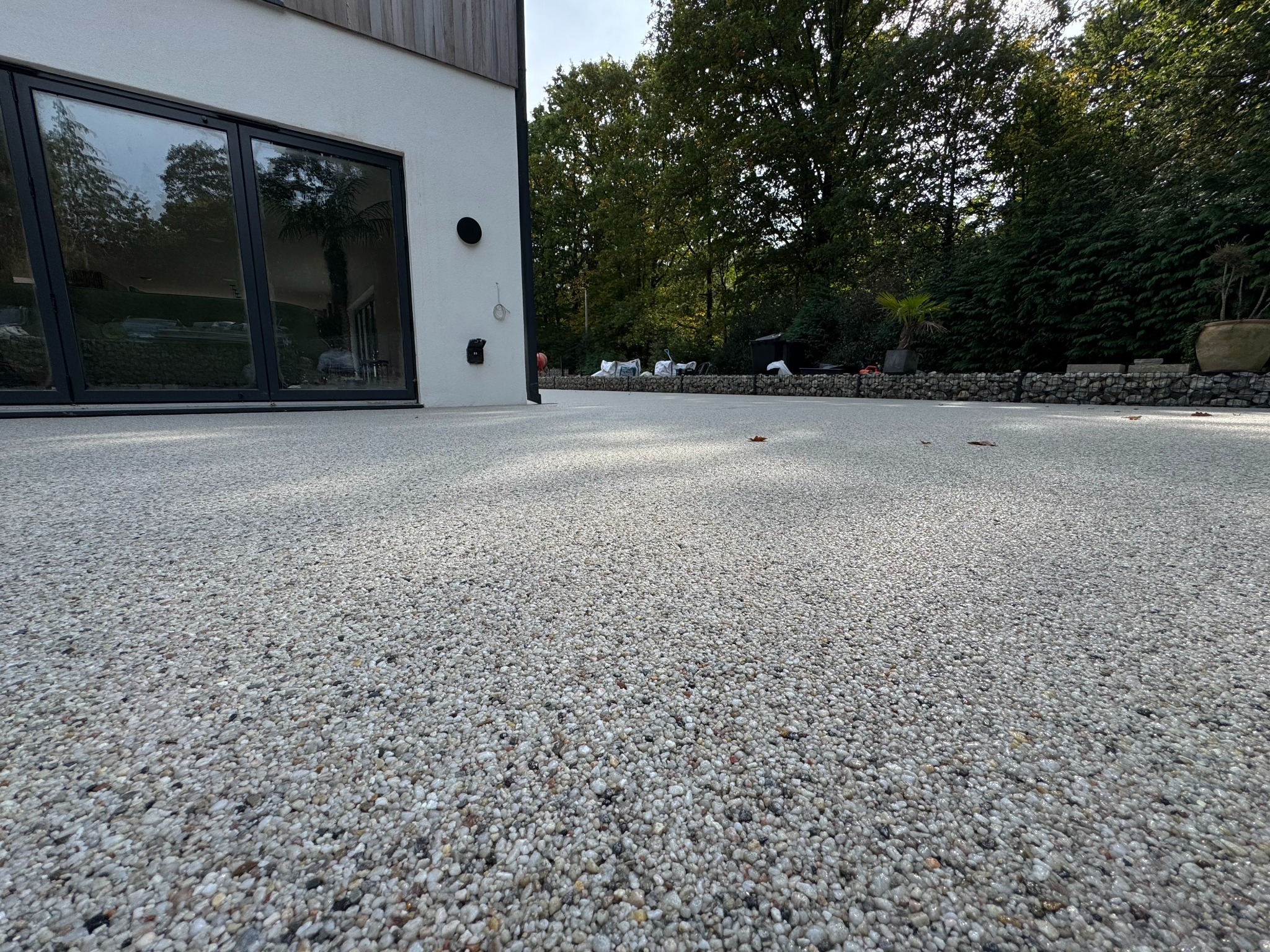 Jasna, kamyczkowa powierzchnia tarasu z żywicy epoksydowej, przylegająca do nowoczesnego domu z dużymi oknami i zielonym ogrodem w tle. Widok z niskiej perspektywy.
