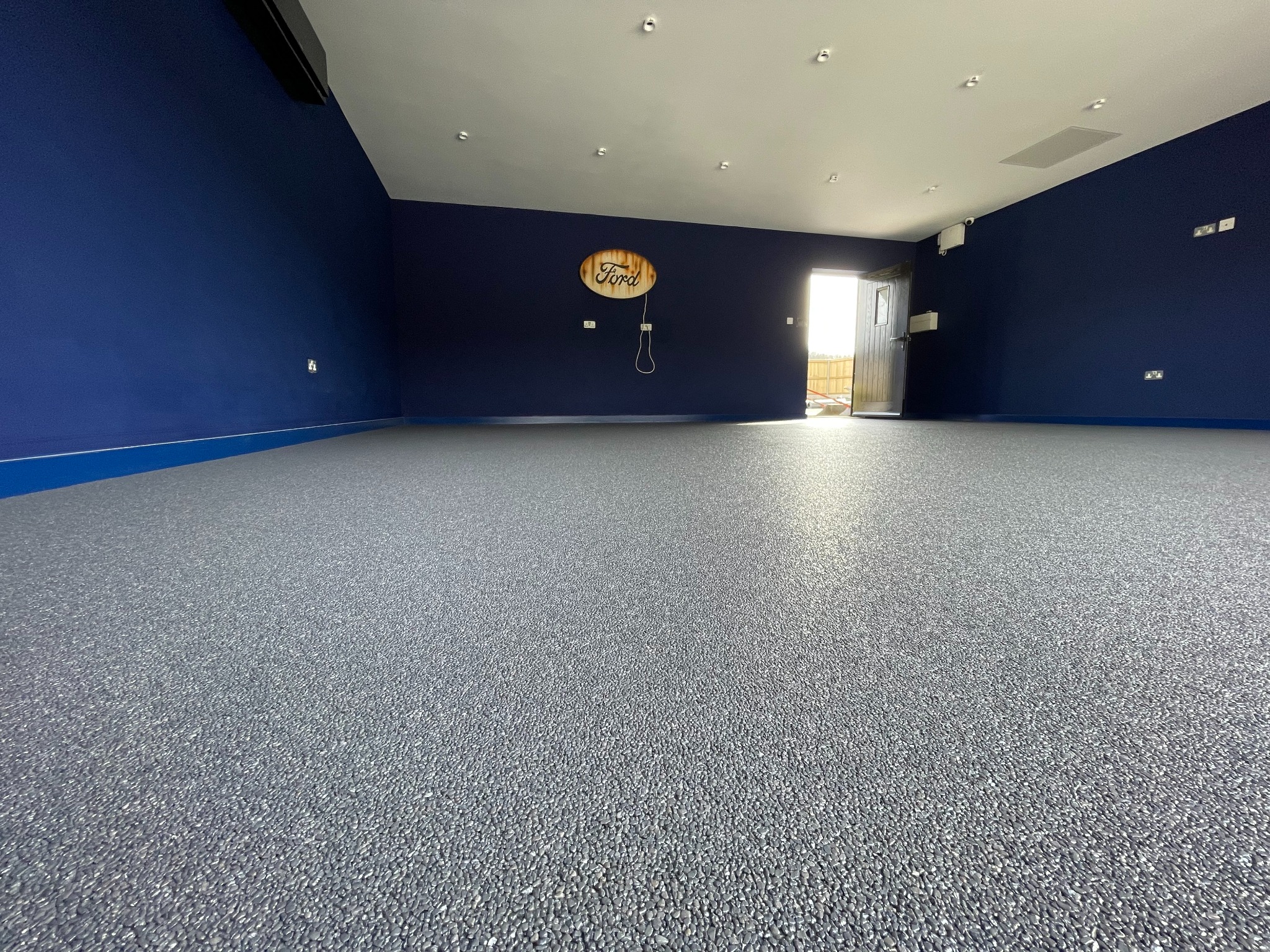 Szara, drobnoziarnista posadzka w garażu z ciemnoniebieskimi ścianami i drewnianym logo Forda. Otwarta brama w tle wpuszcza jasne światło.