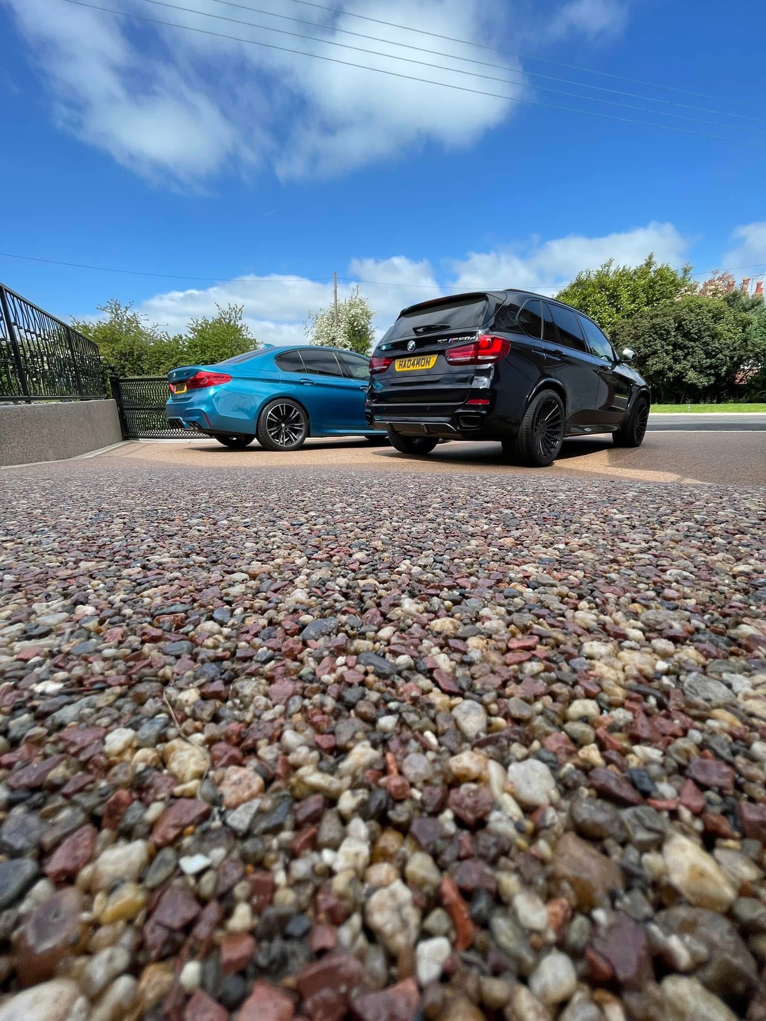 Żwirowa nawierzchnia podjazdu z zaparkowanymi samochodami BMW w kolorze niebieskim i czarnym. Widok z niskiej perspektywy, błękitne niebo w tle.