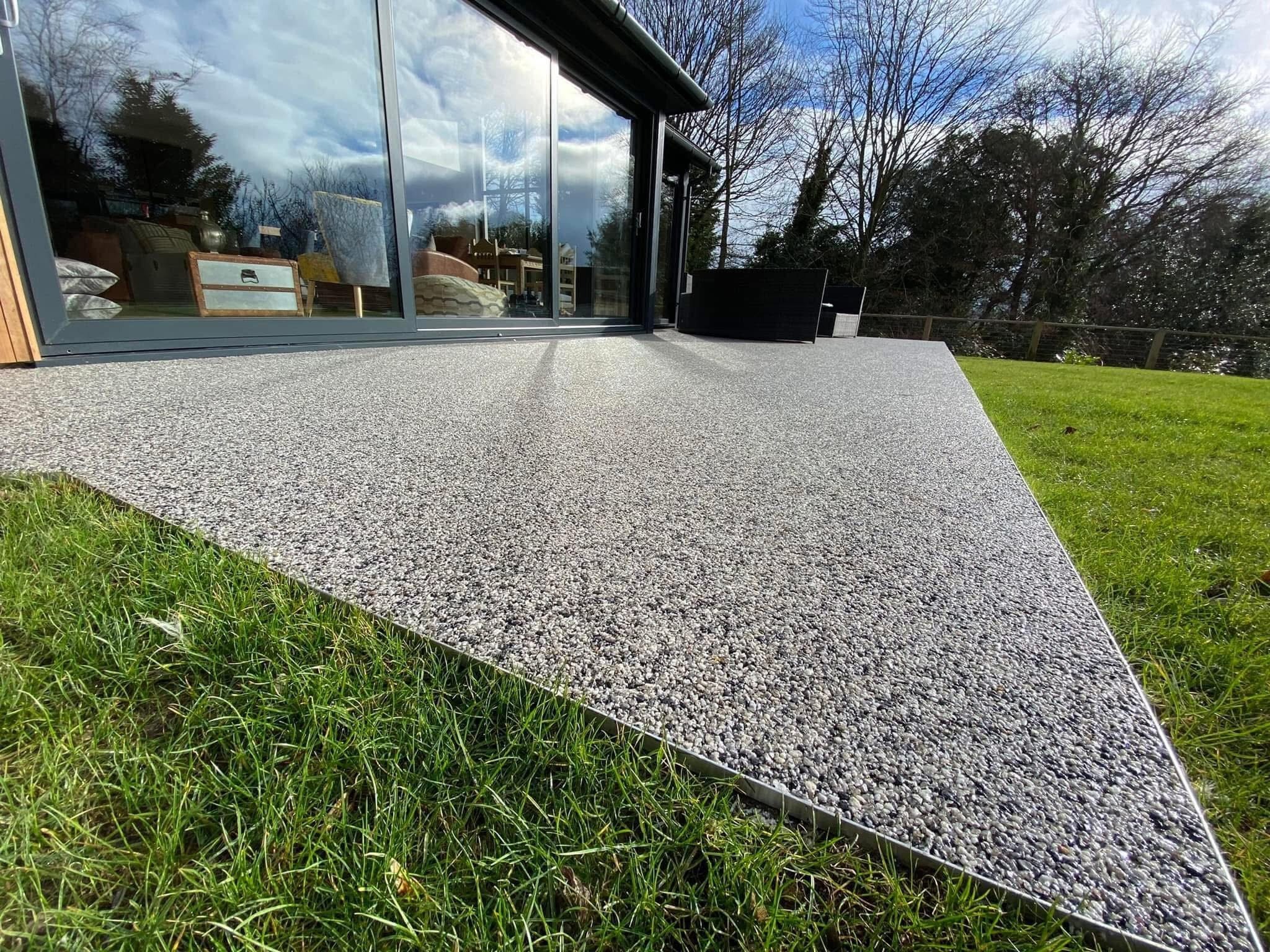 Szara, kamyczkowa nawierzchnia tarasu z metalowym obrzeżem, przylegająca do trawnika i przeszklonego budynku w tle, w słoneczny dzień.