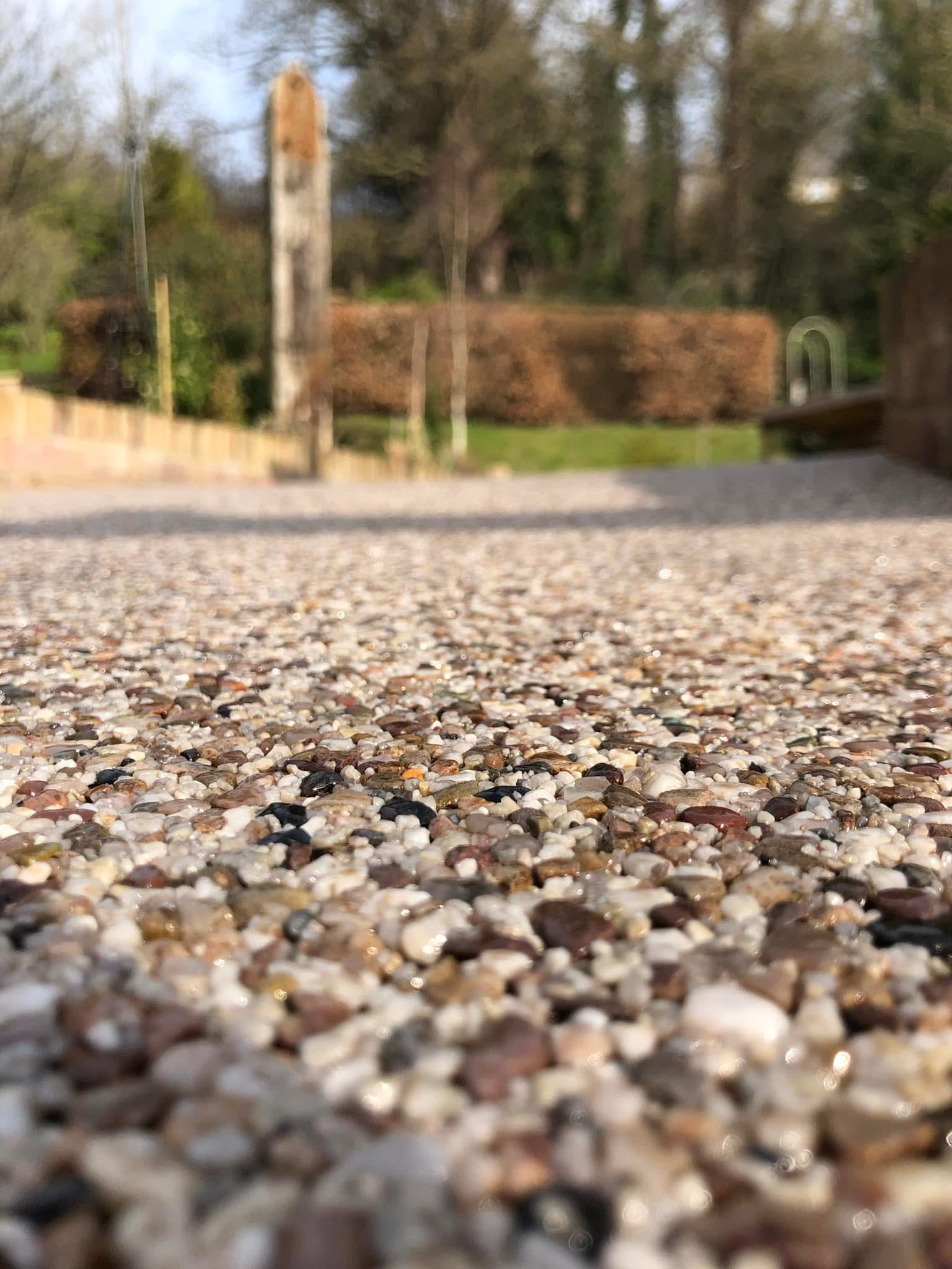 Żwirowa nawierzchnia z żywicy epoksydowej w ogrodzie, z widocznym tłem zieleni i elementami architektury ogrodowej. Niska perspektywa podkreśla fakturę.