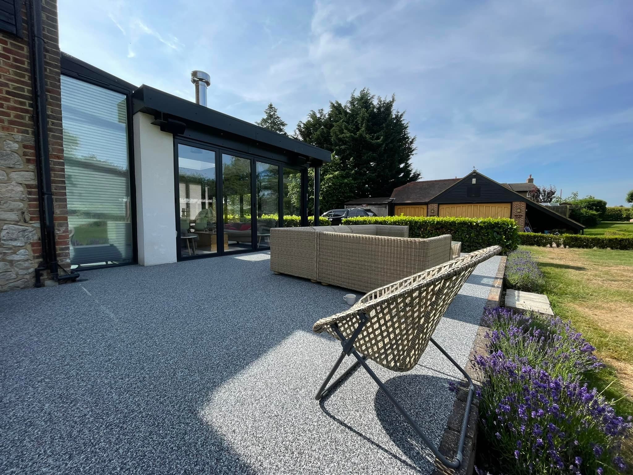 Szara, drobnoziarnista nawierzchnia tarasu z żywicy epoksydowej, z wiklinowym fotelem i sofą. W tle nowoczesny dom z dużymi oknami i zadbany ogród.