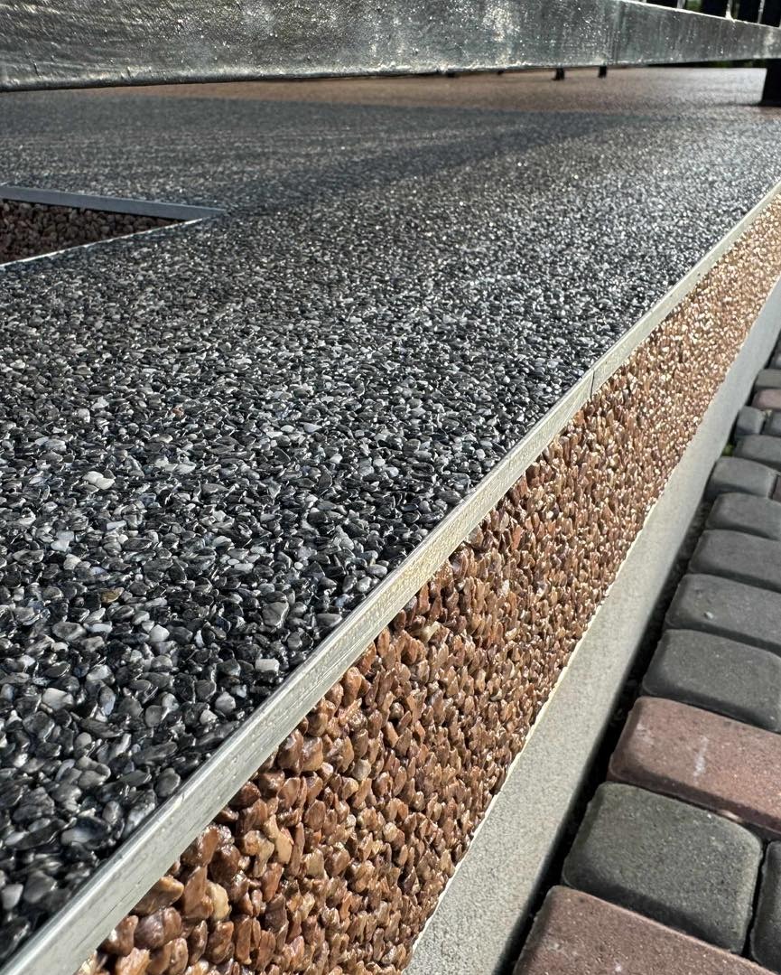 Żywiczna posadzka z drobnych kamyczków w odcieniach szarości i brązu, oddzielona metalową listwą. Widoczny fragment chodnika z kostki brukowej.