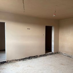 Usługi Ogólnobudowlane Janusz Lewandowski - Wnętrze po tynkowaniu maszynowym: gładkie, beżowe ściany, widoczne kable wystające z sufitu, przygotowane do dalszych prac wykończeniowych. Surowy stan podłogi.