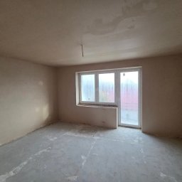 Usługi Ogólnobudowlane Janusz Lewandowski - Wnętrze pokoju po nałożeniu tynków: gładkie, jasne ściany i sufit, okno z widokiem na zewnątrz. Podłoga surowa, przygotowana do dalszych prac wykończeniowych.