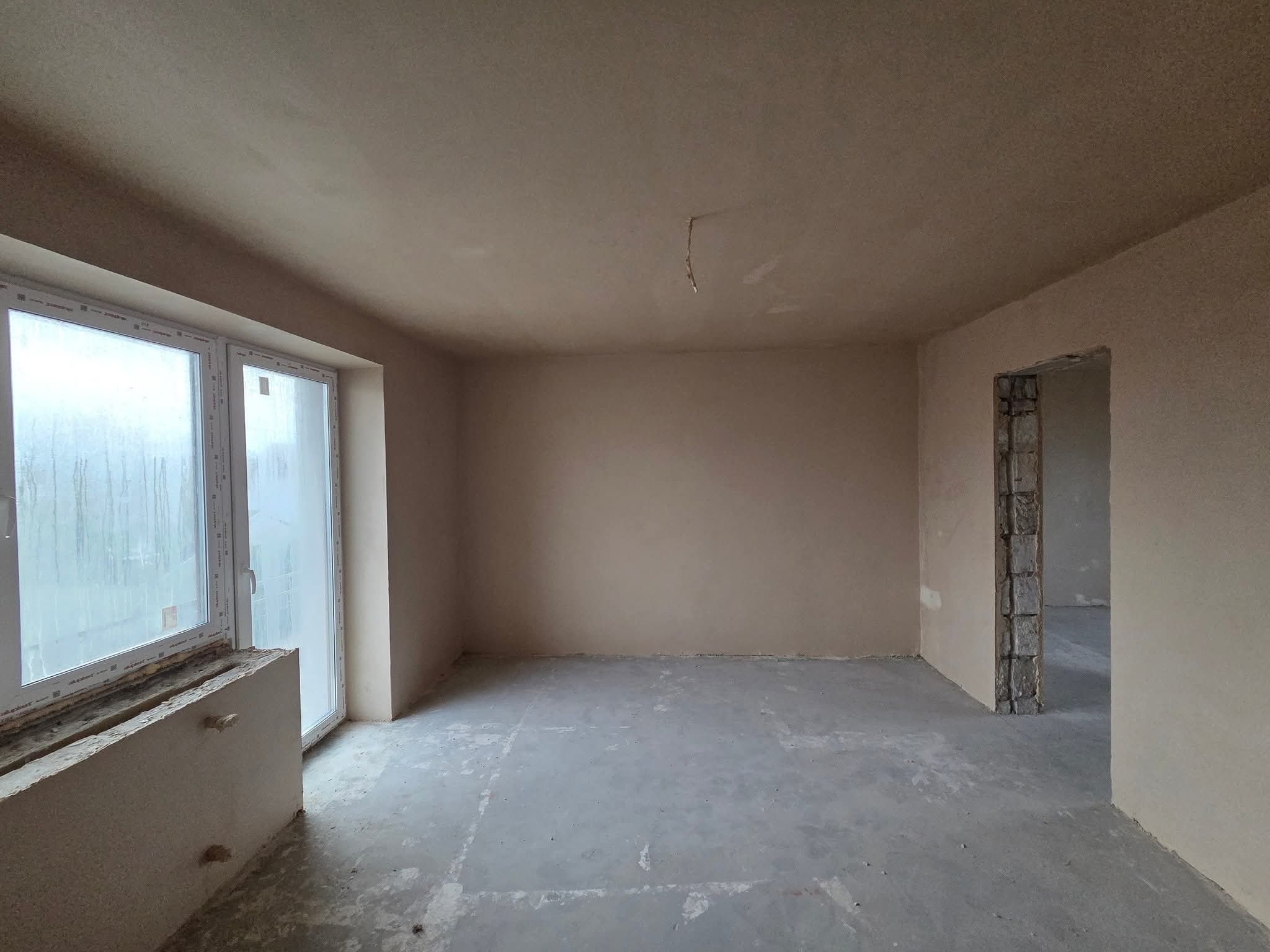 Wnętrze po tynkowaniu maszynowym: gładkie, jasne ściany, surowy beton na podłodze, okno z widocznymi śladami wilgoci, surowy otwór drzwiowy. Minimalistyczny efekt.