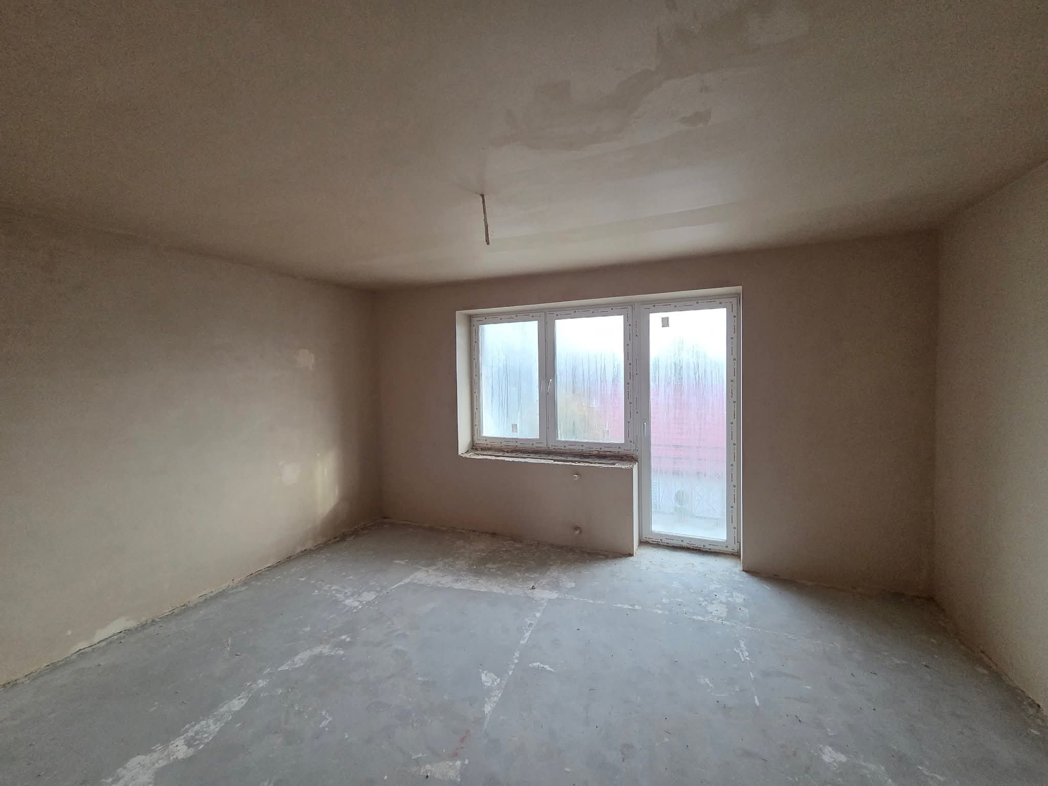 Pokój po nałożeniu tynków: gładkie ściany w odcieniach beżu, surowa podłoga, okno z widokiem na zewnątrz. Widoczne niedoskonałości i surowość wykończenia.