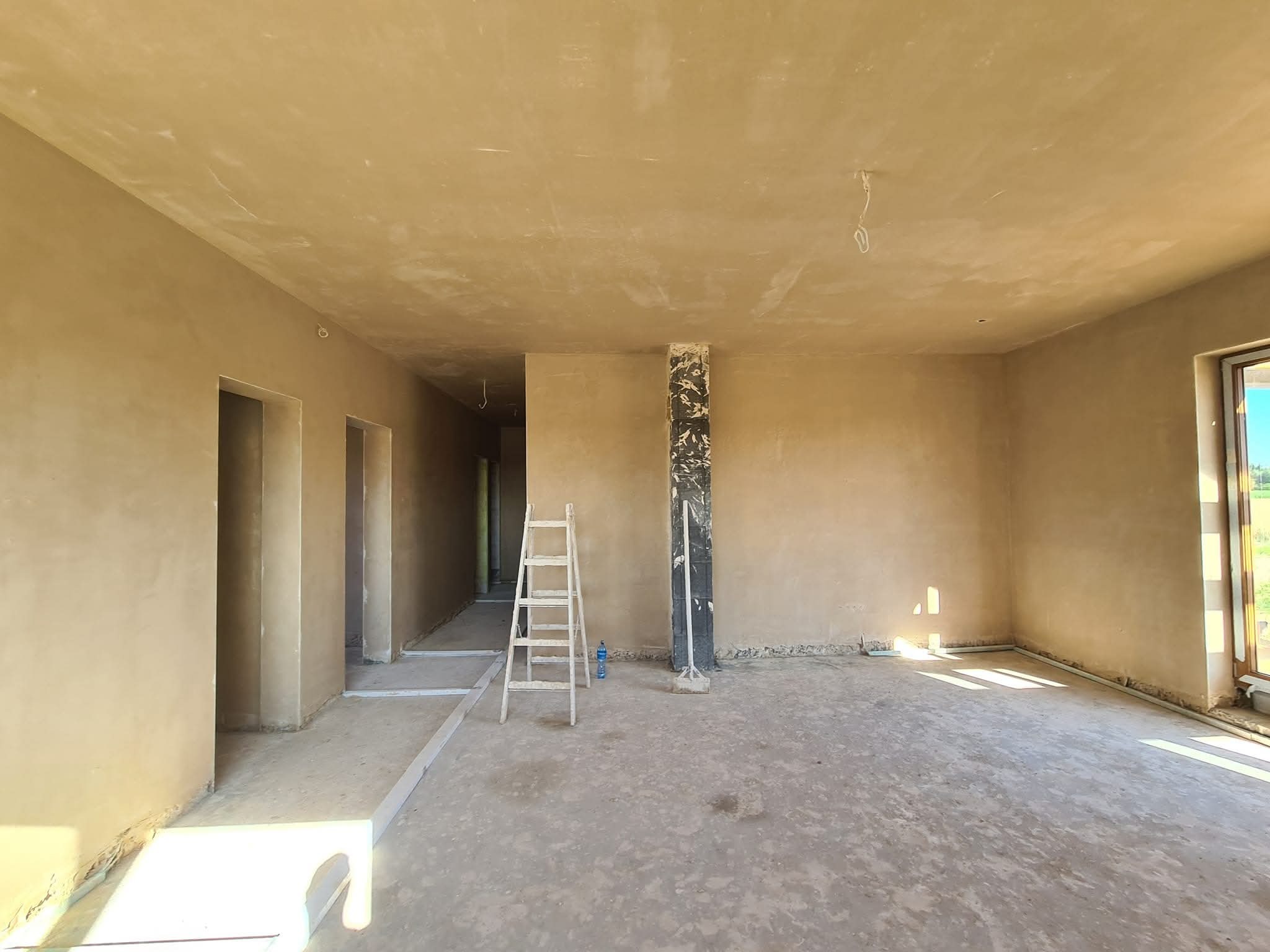 Wnętrze po tynkowaniu maszynowym: jasne ściany, surowy beton na podłodze, drabina, zabezpieczona kolumna. Widoczne ślady po pracach tynkarskich.