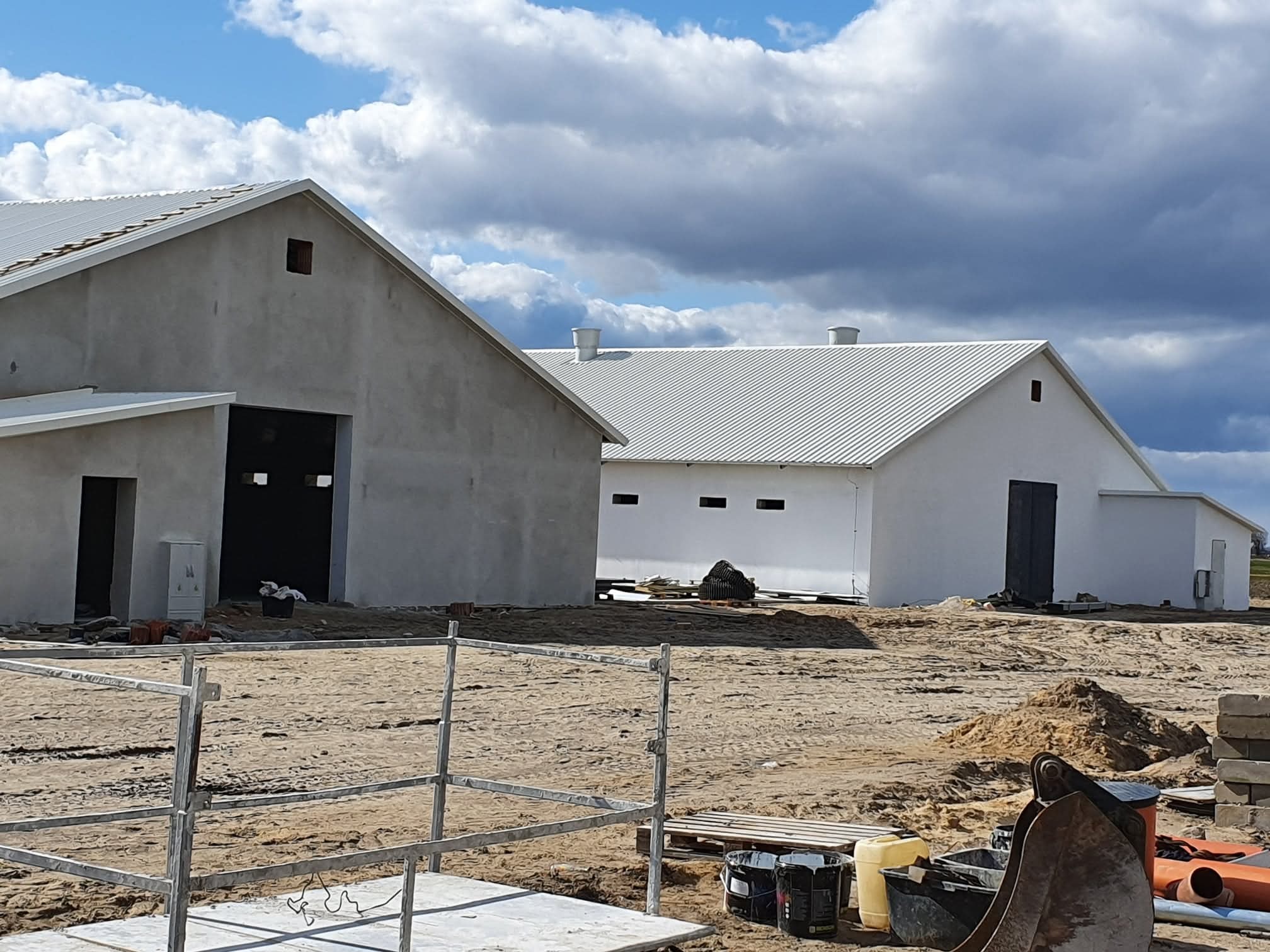 Dwa budynki gospodarcze z szarymi ścianami i białymi dachami blaszakami, w trakcie budowy, na piaszczystym terenie z rusztowaniami i sprzętem budowlanym.