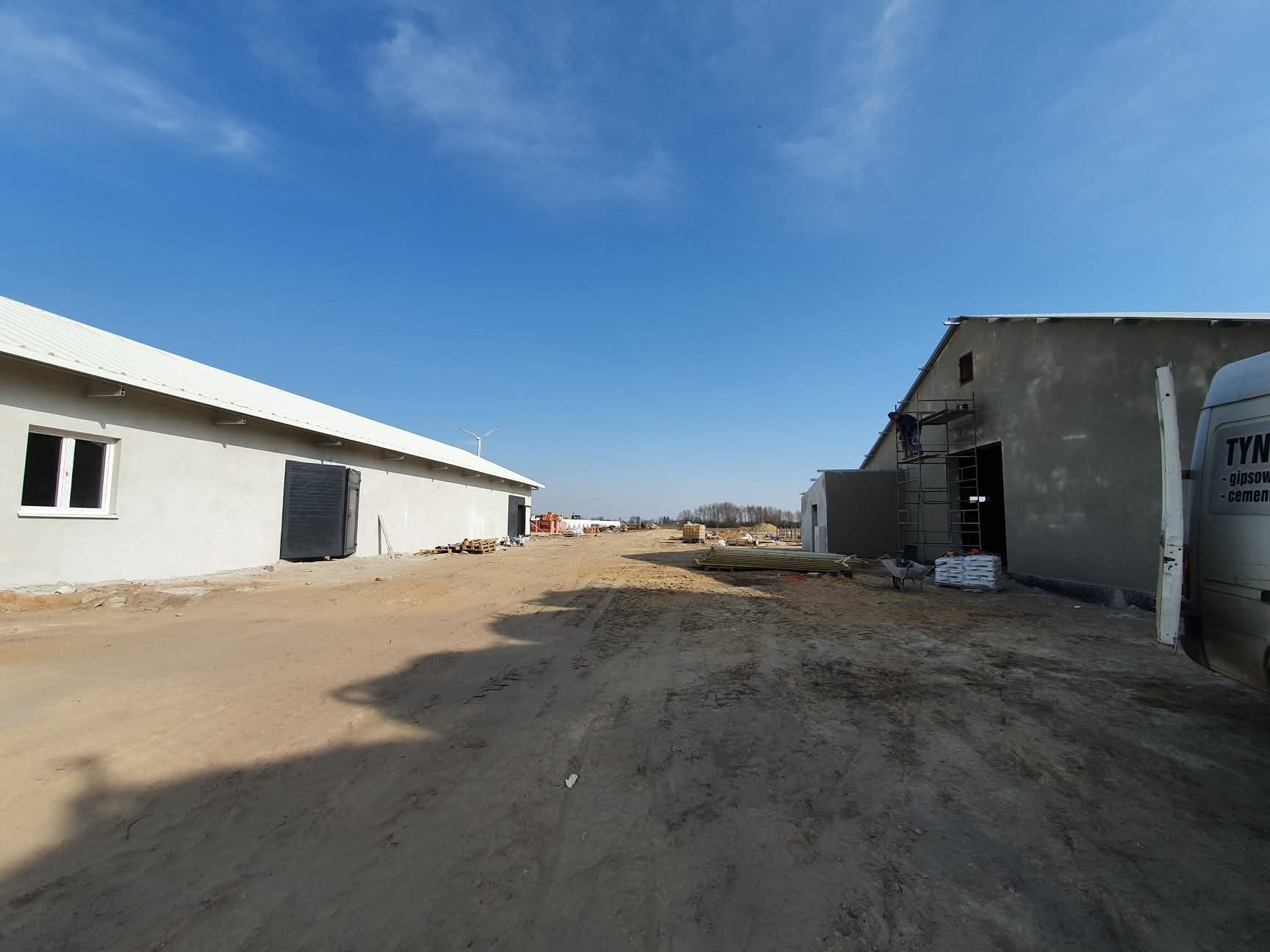 Tynkowanie maszynowe budynku gospodarczego w trakcie realizacji. Widoczny pracownik na rusztowaniu, worki z zaprawą i samochód dostawczy z logo firmy.