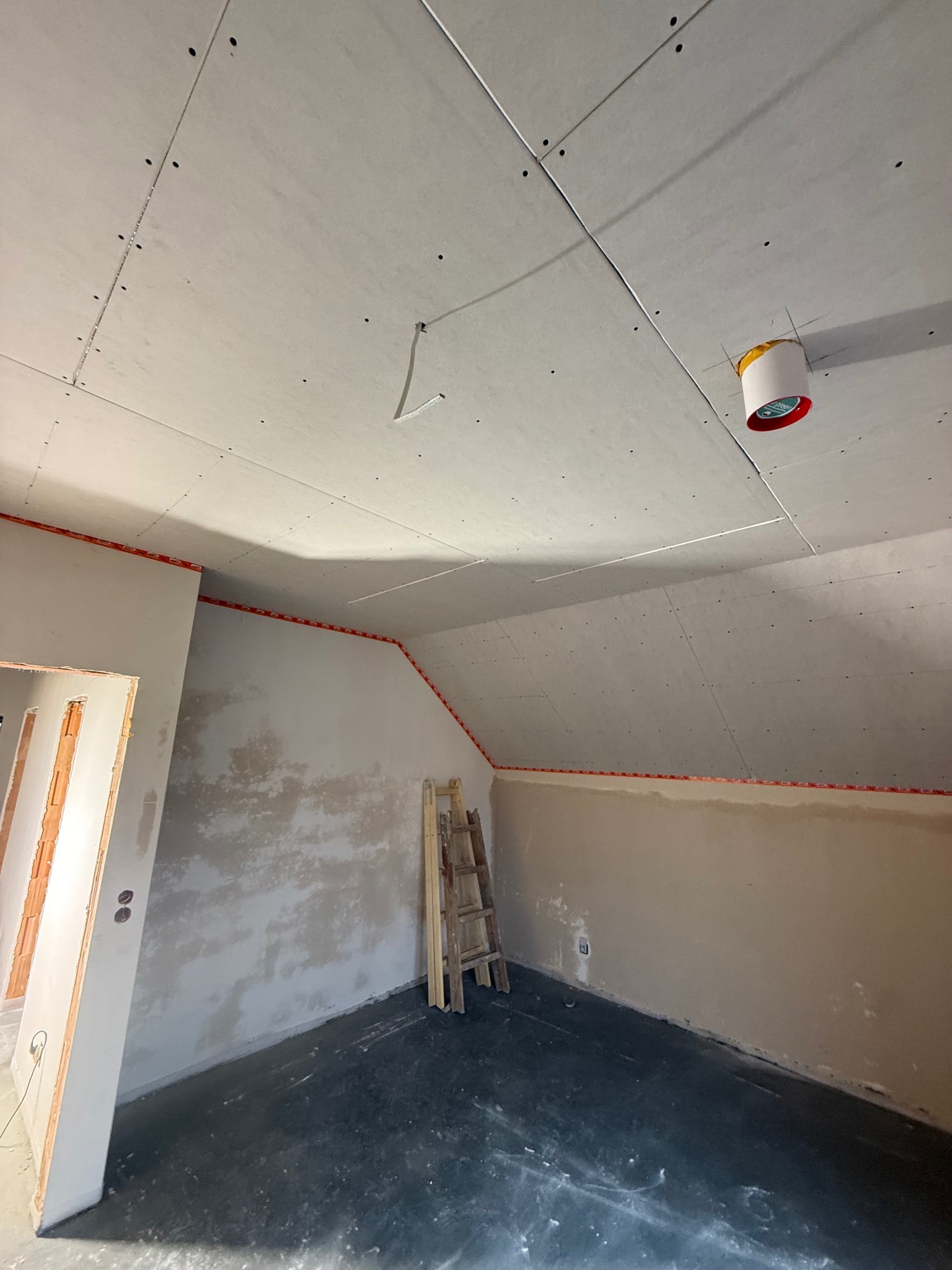 Poddasze w trakcie prac wykończeniowych: zamontowane płyty karton-gips na suficie, ściany przygotowane do malowania, widoczna drabina. Oświetlenie tymczasowe.