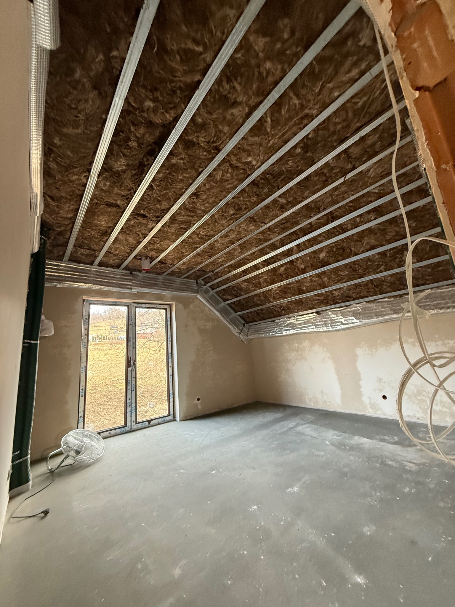 Poddasze w trakcie adaptacji: widoczne ocieplenie wełną mineralną między metalowymi profilami sufitowymi, surowe ściany i wylewka podłogowa, okno z widokiem na zewnątrz.