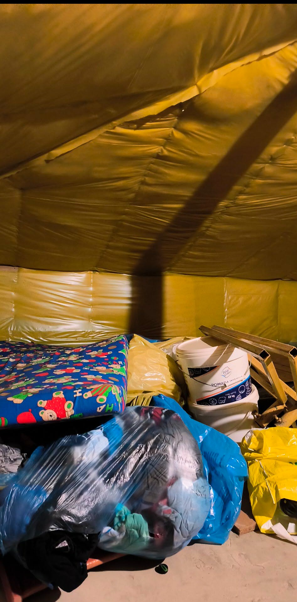 Wnętrze pomieszczenia z żółtą folią na ścianach, wypełnione workami, wiadrem z farbą i materacem dziecięcym. Przygotowanie do opróżniania mieszkania.