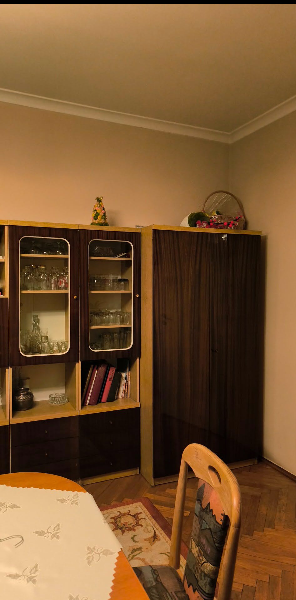 Masywna, drewniana meblościanka z przeszklonymi witrynami, wypełnionymi szkłem i książkami. Na górze dekoracje. Część stołu i krzesła w kadrze. Parkiet na podłodze.